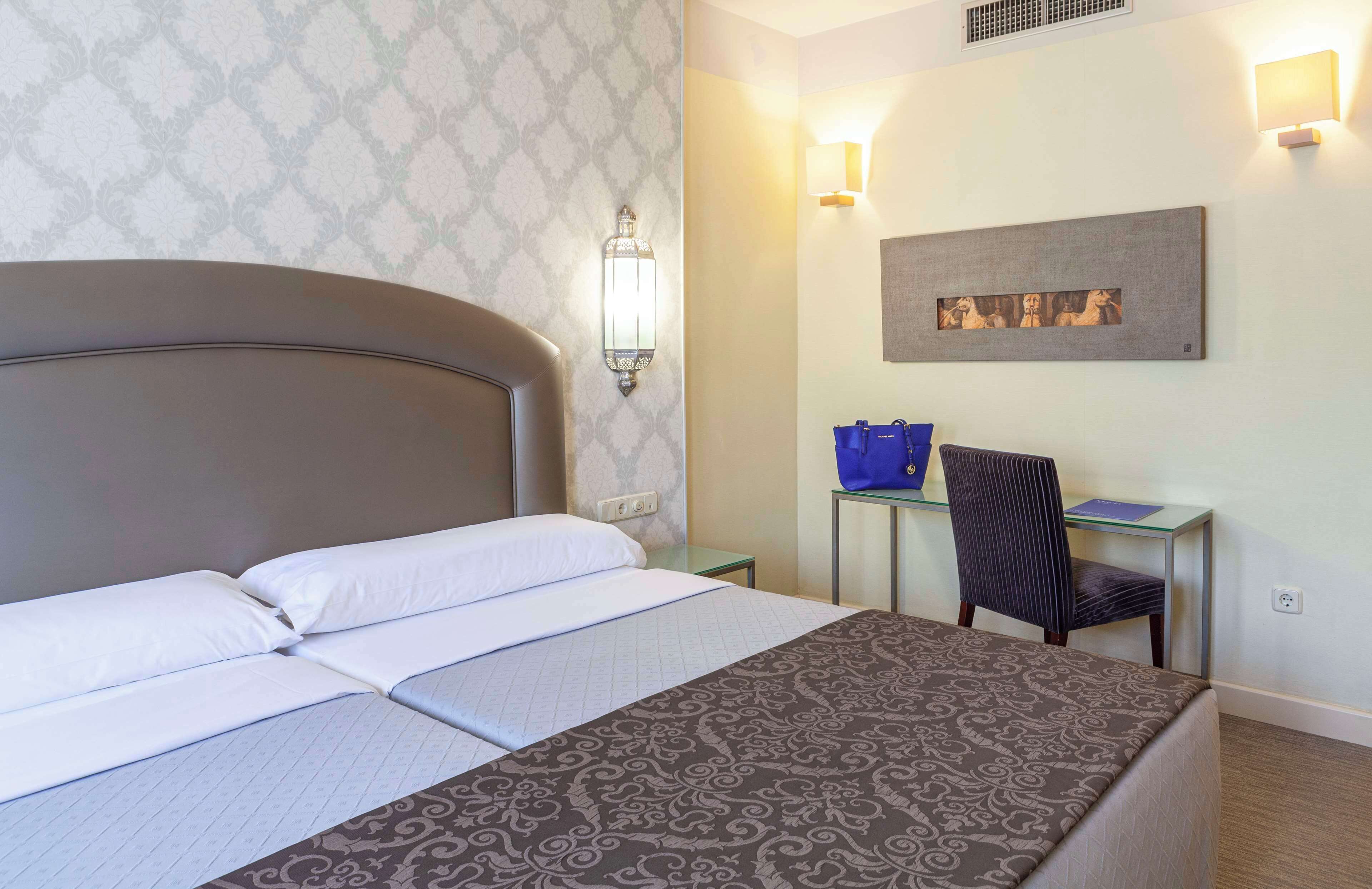 A hotel room with a large bed, a desk with a chair, and a wall-mounted light fixture.