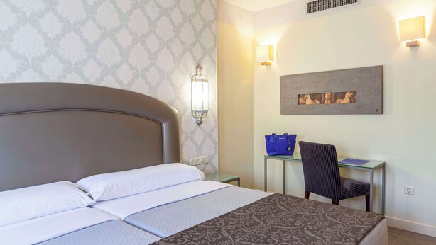 A hotel room with a large bed, a desk with a chair, and a wall-mounted light fixture. - Hotel Macià Alfaros (Córdoba)