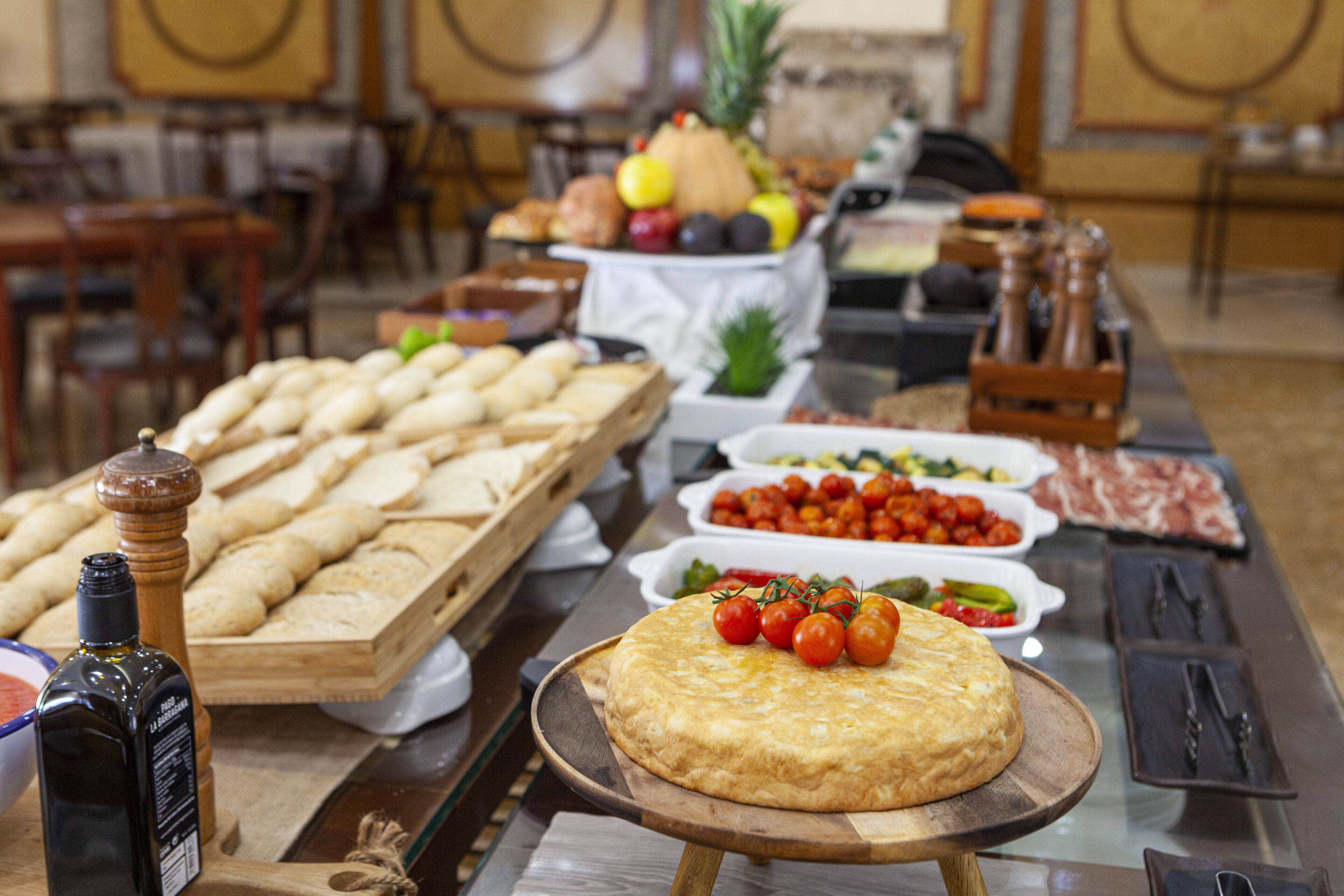 A buffet table with various food items including bread, a cheese plate with tomatoes, and a bottle of olive oil.
