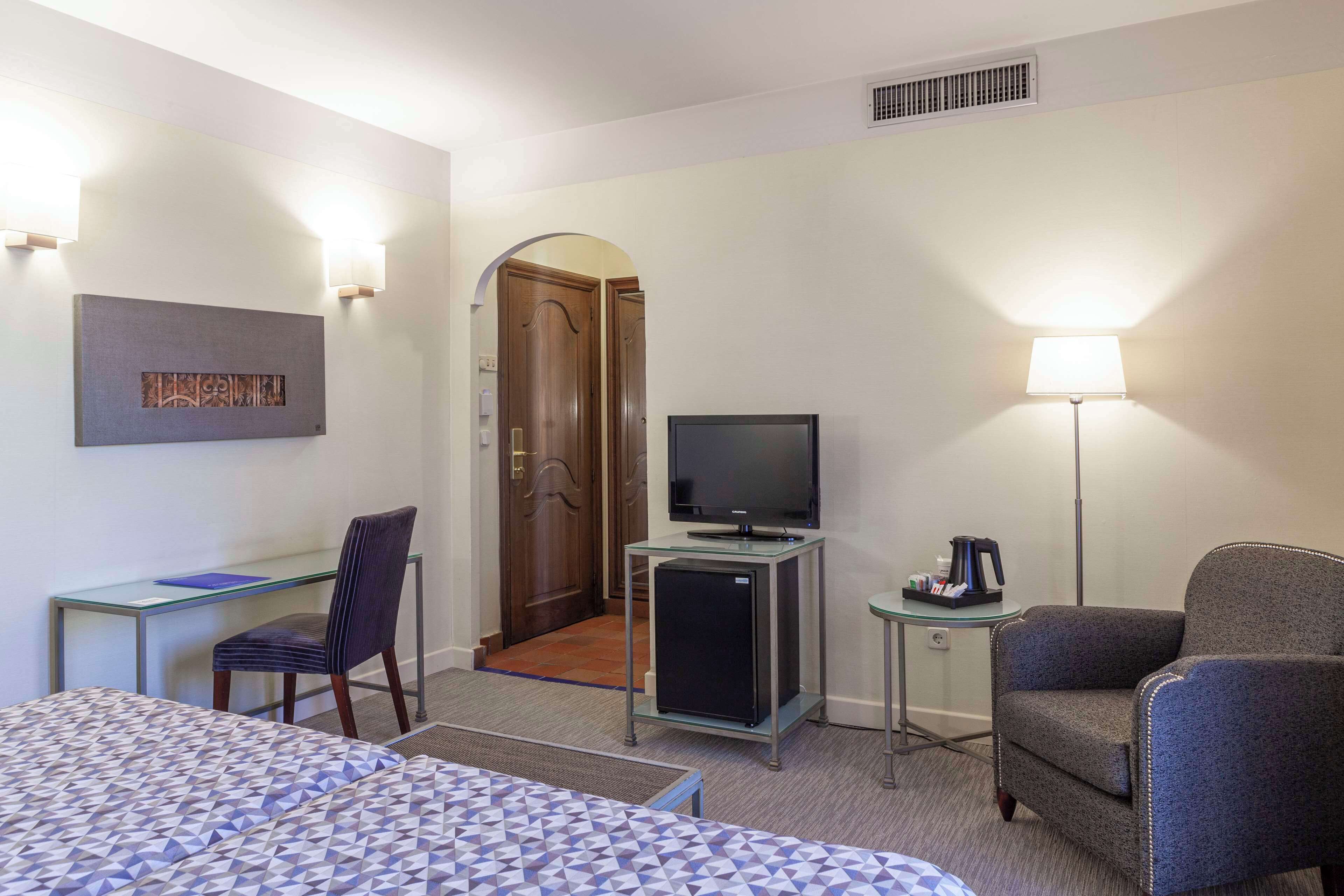 A hotel room with a bed, a desk with a chair, a television, a refrigerator, a lamp, and a door leading to another room.