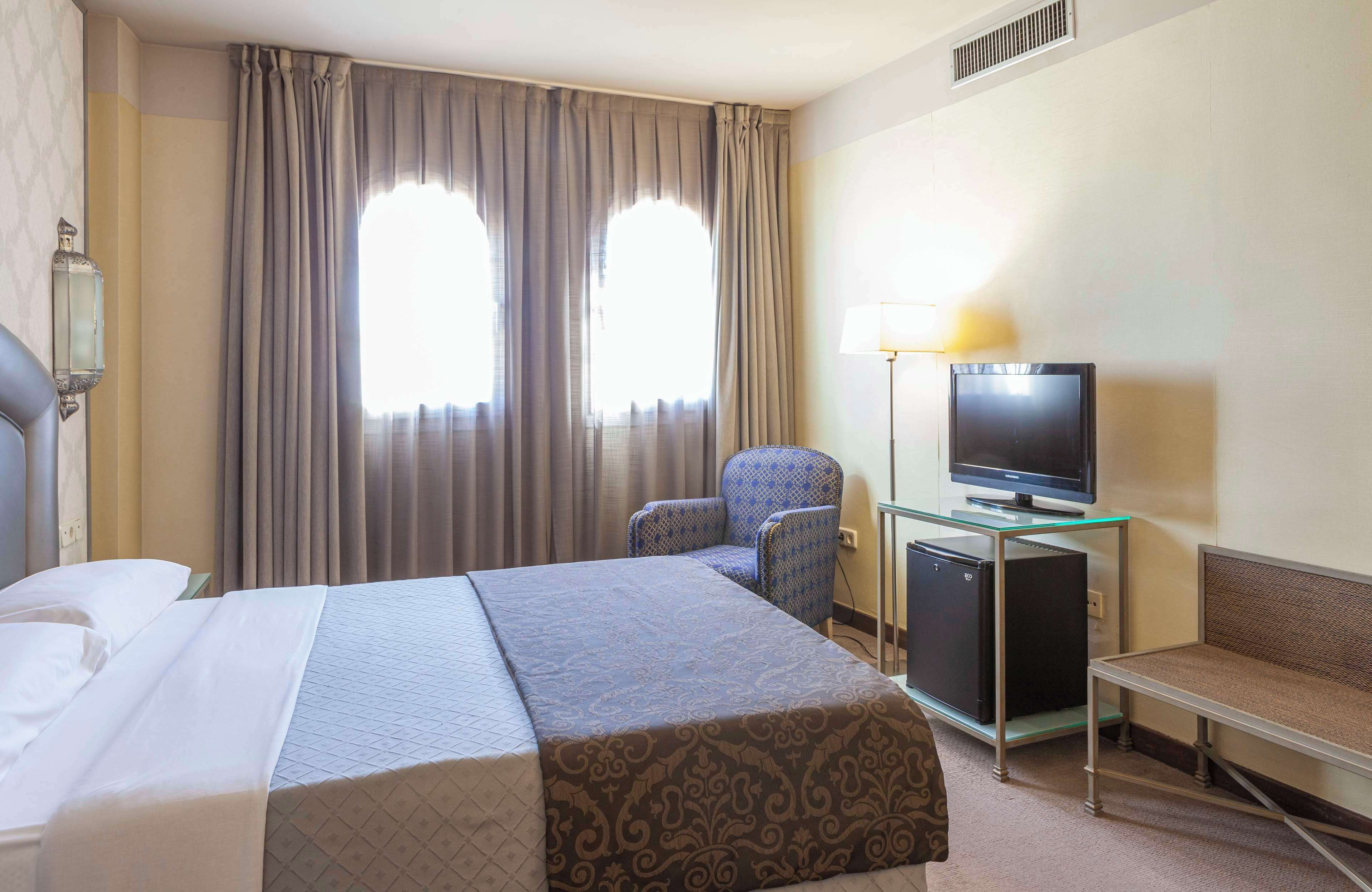 A hotel room with a bed, a desk with a television, a chair, and a lamp.