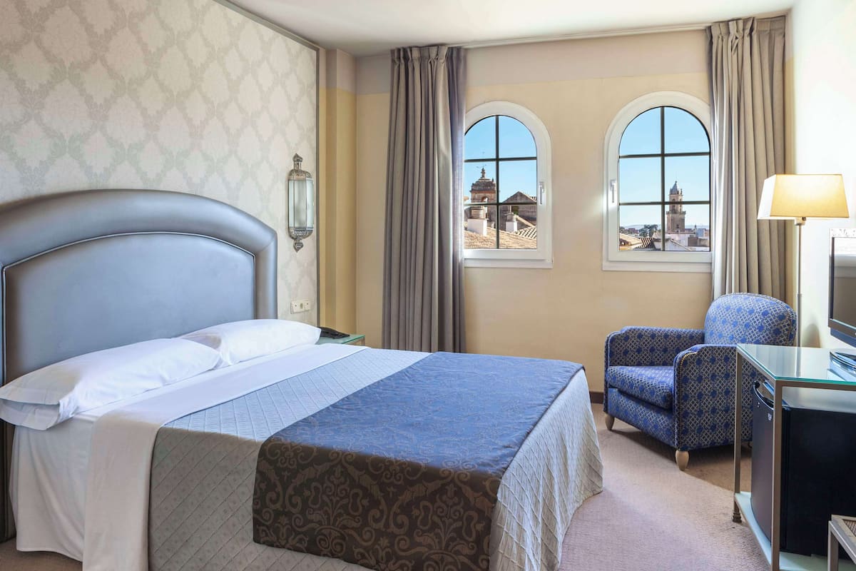 Habitación de hotel con una cama grande, dos ventanas que dan a un edificio histórico, un escritorio con lámpara y una silla.