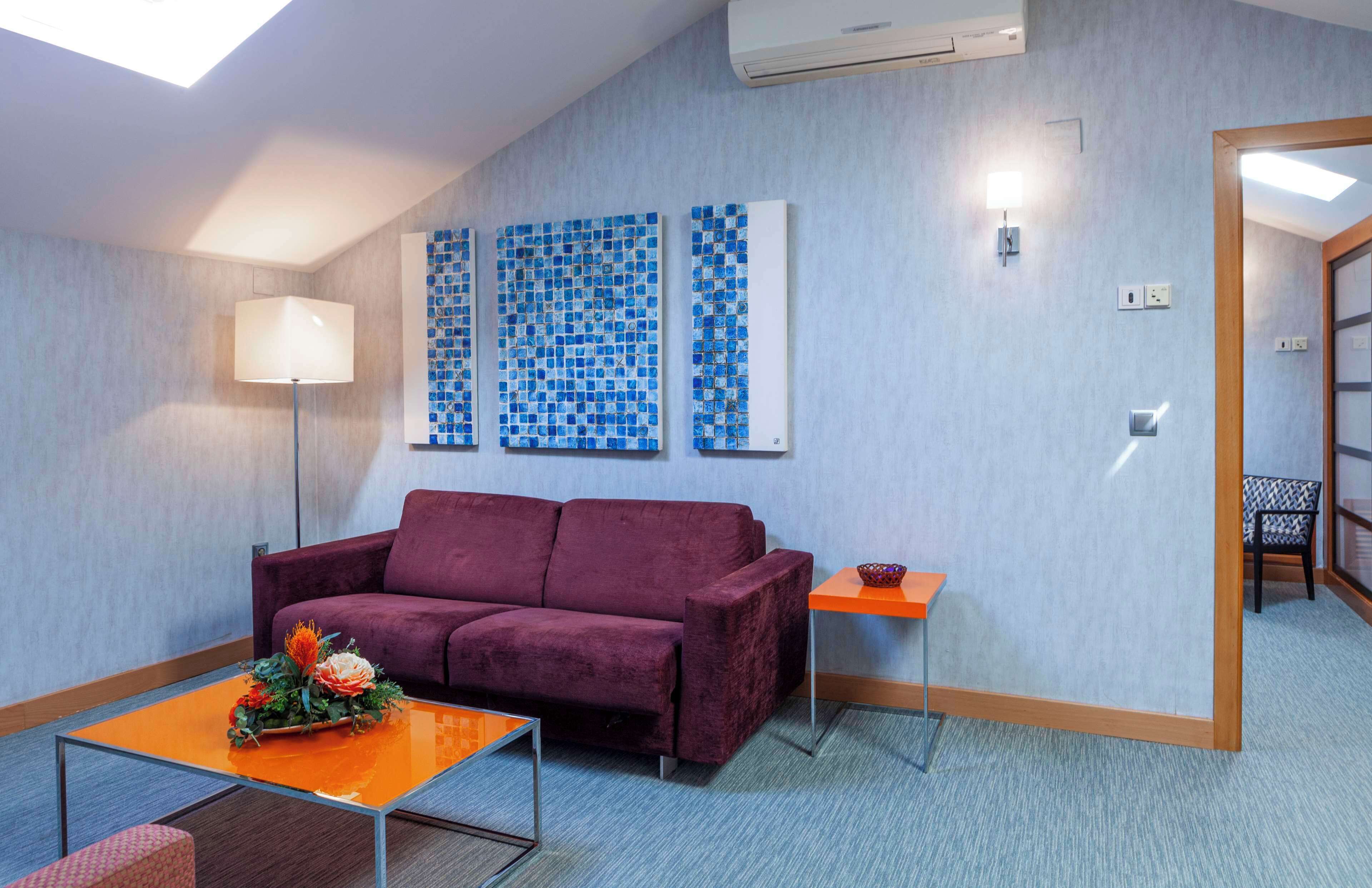 A modern room with a purple sofa, a small orange table with a vase, and blue mosaic art on the wall.