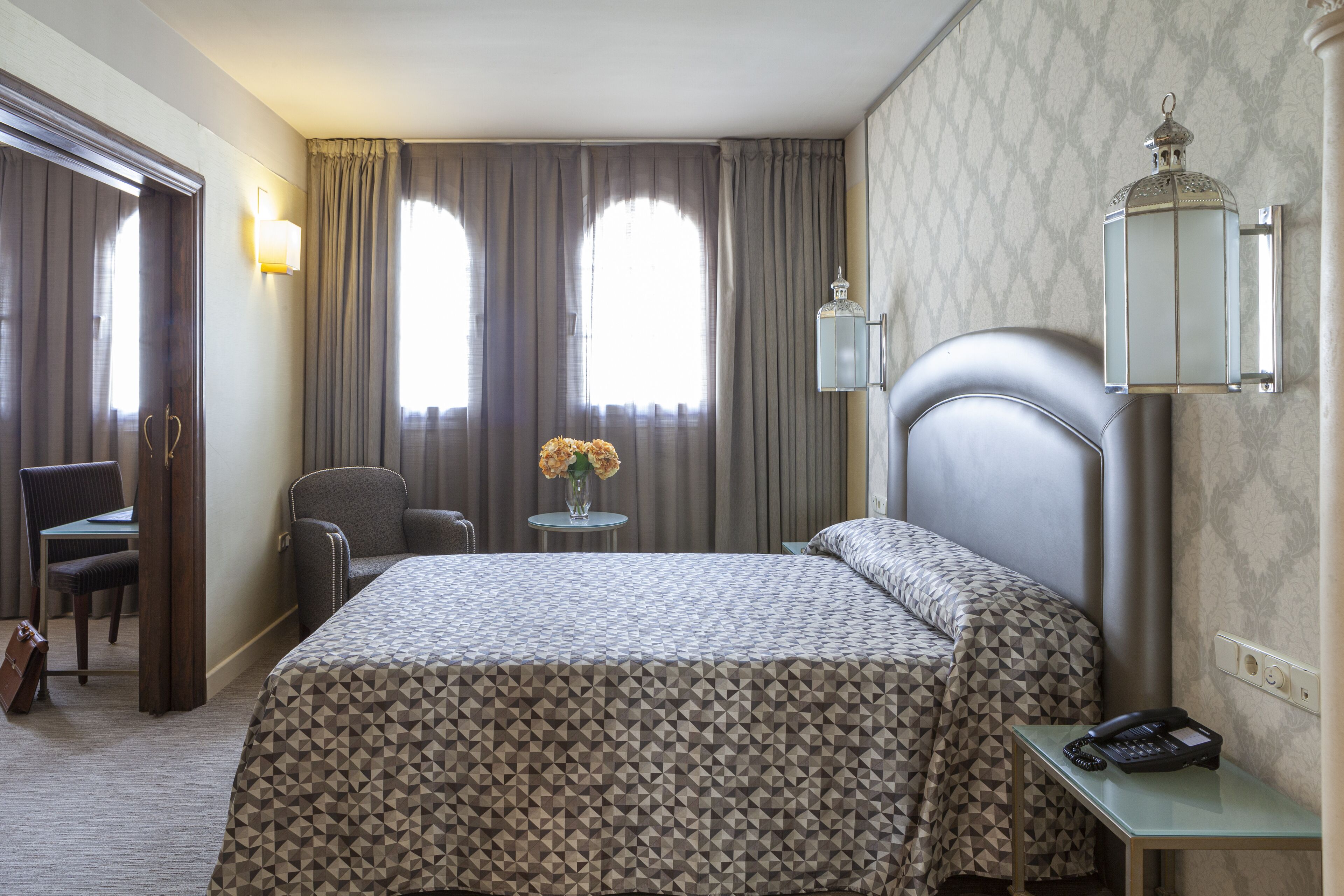 Una camera d'albergo con un letto, una scrivania, una sedia, una lampada e un vaso con fiori.