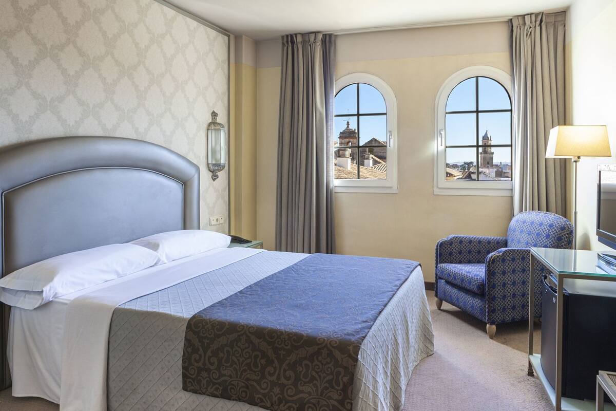 A hotel room with a bed, a desk with a computer, a chair, and two windows with a view of buildings.