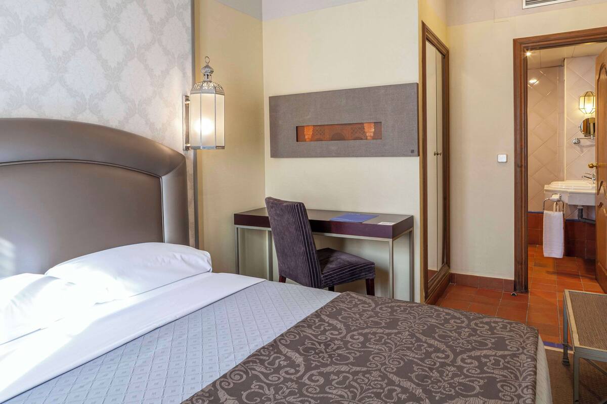 Habitación de hotel con cama, escritorio, silla y baño visible a través de una puerta abierta.