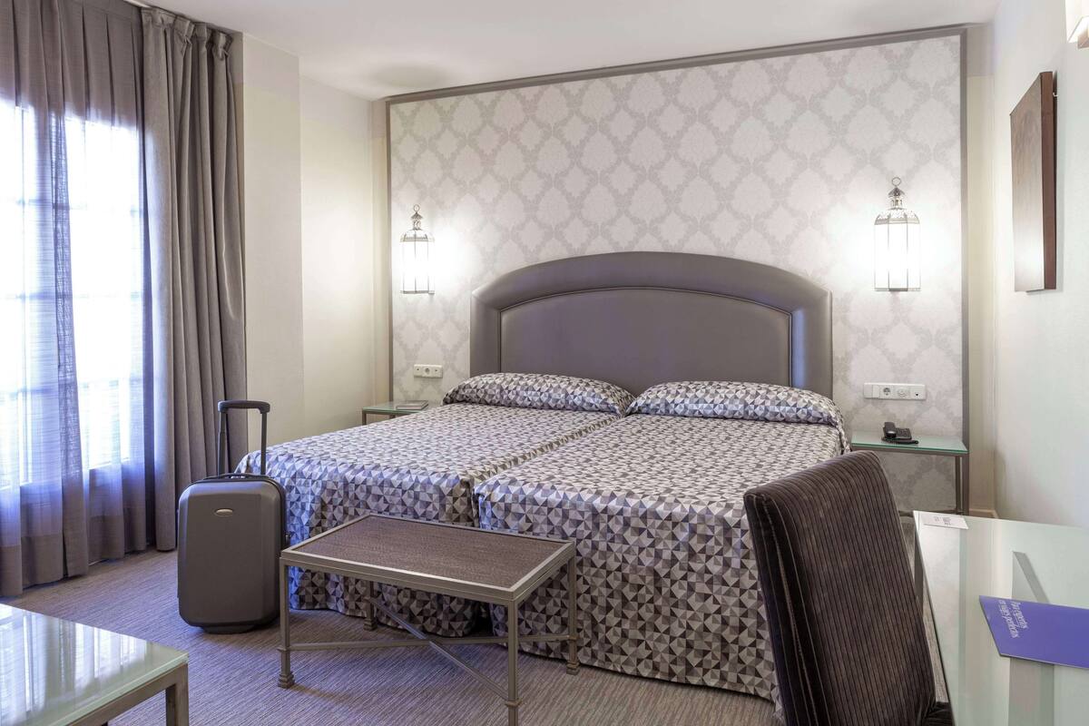 Una habitación de hotel con dos camas, un escritorio, una silla, una maleta y una lámpara.