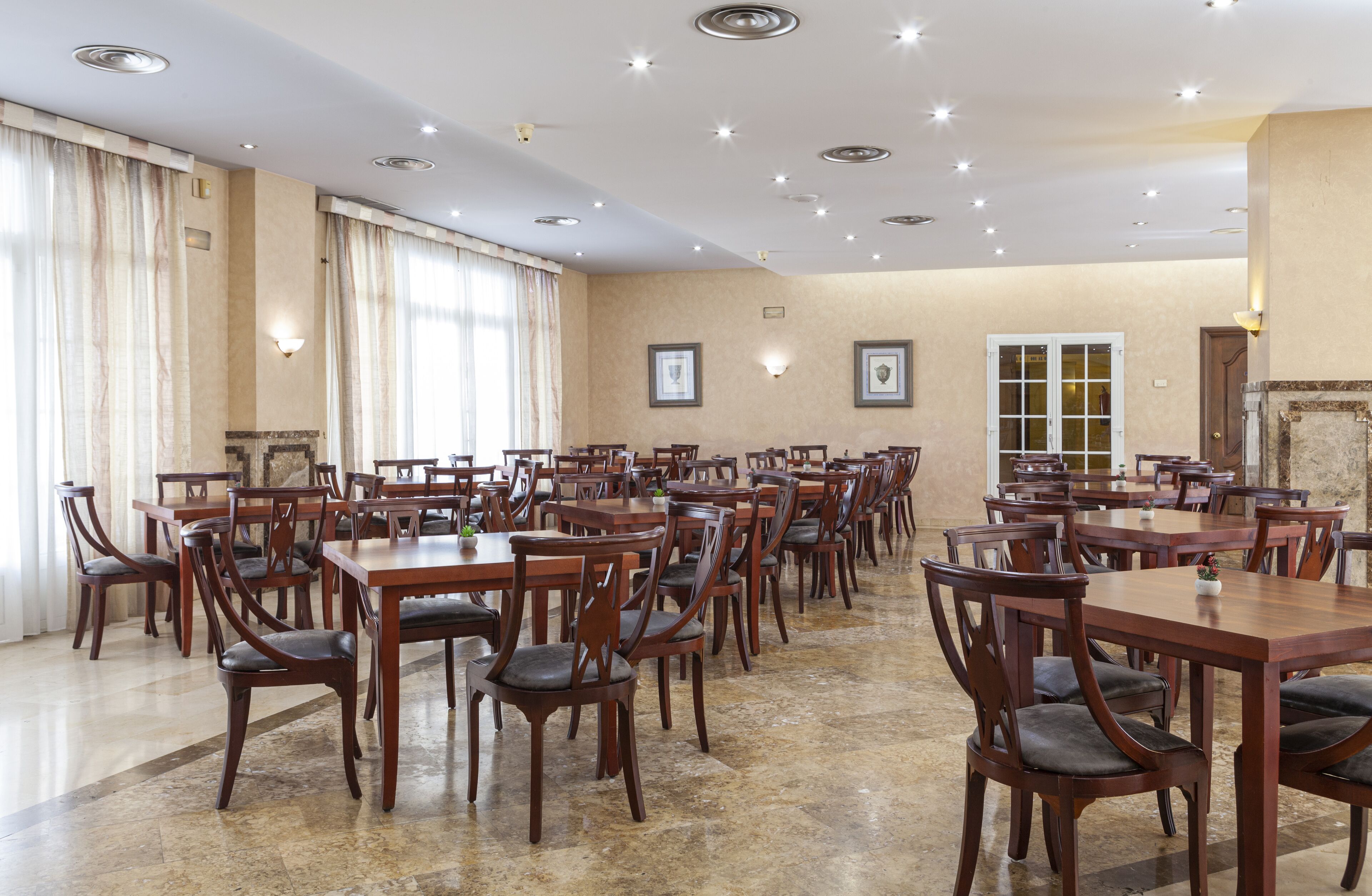 A spacious dining area with multiple wooden tables and chairs, recessed ceiling lights, and framed artwork on the walls.
