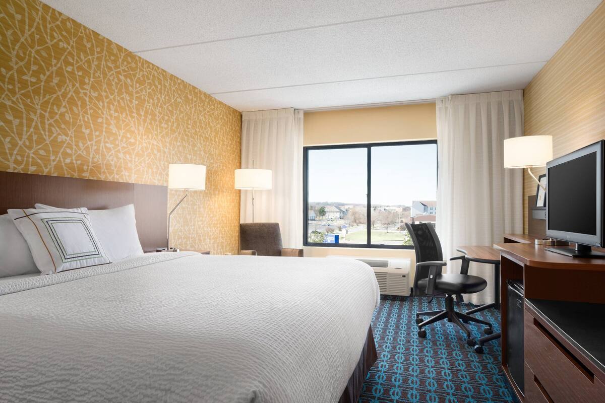 A hotel room with a large bed, a desk with a television, and a view of the city through the window.