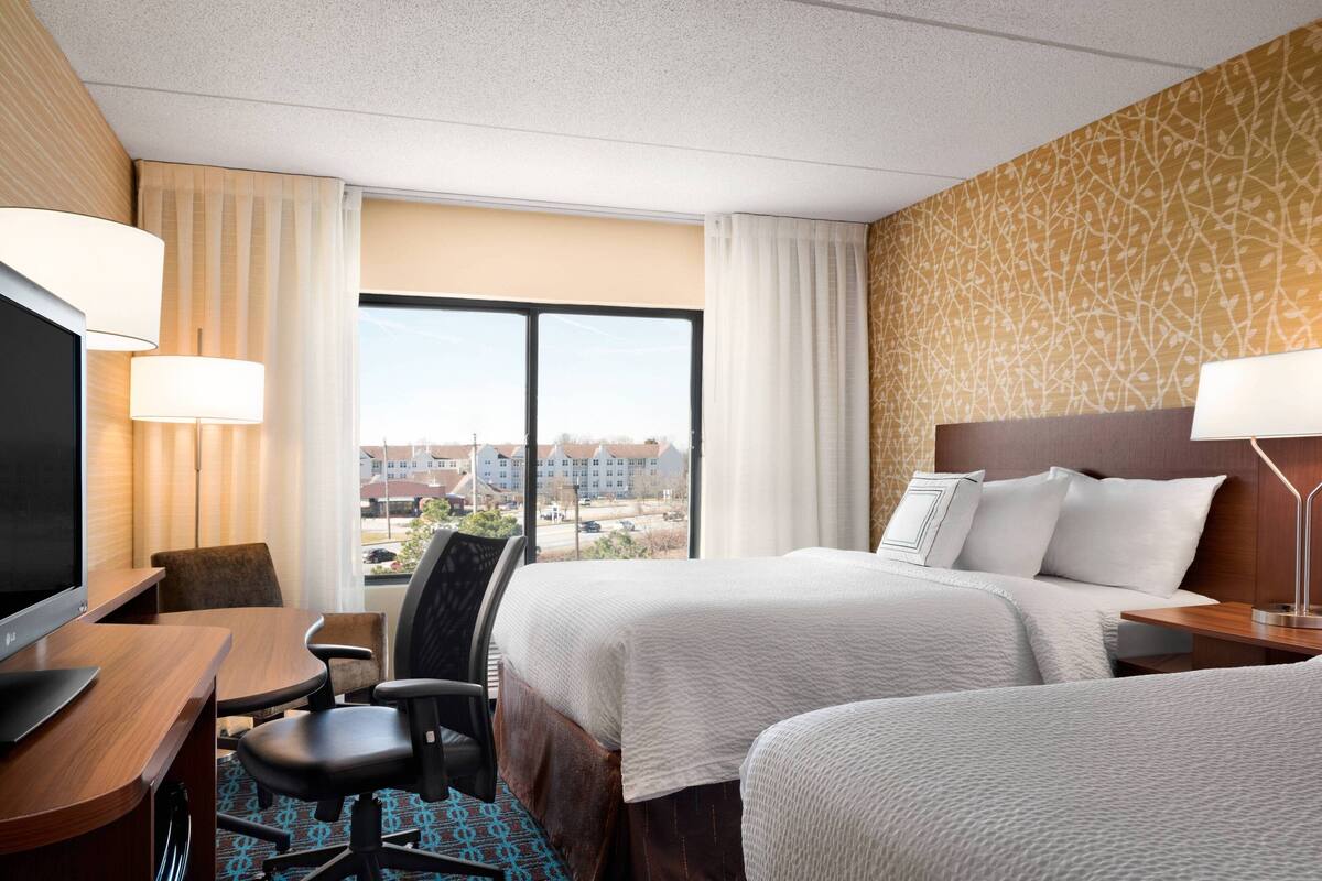A hotel room with two beds, a desk with a TV, and a view of buildings through large windows.