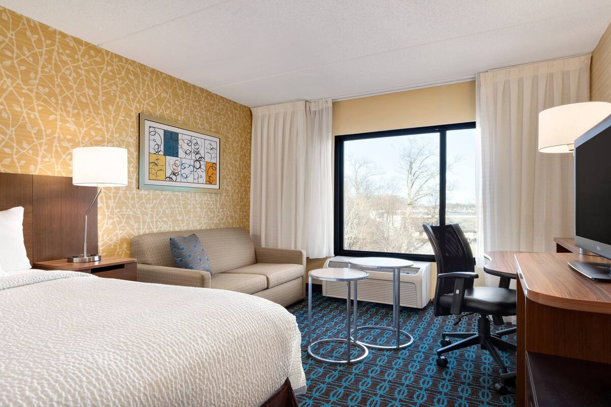 A hotel room with a bed, a sofa, a desk, a TV, a lamp, and a window with curtains.