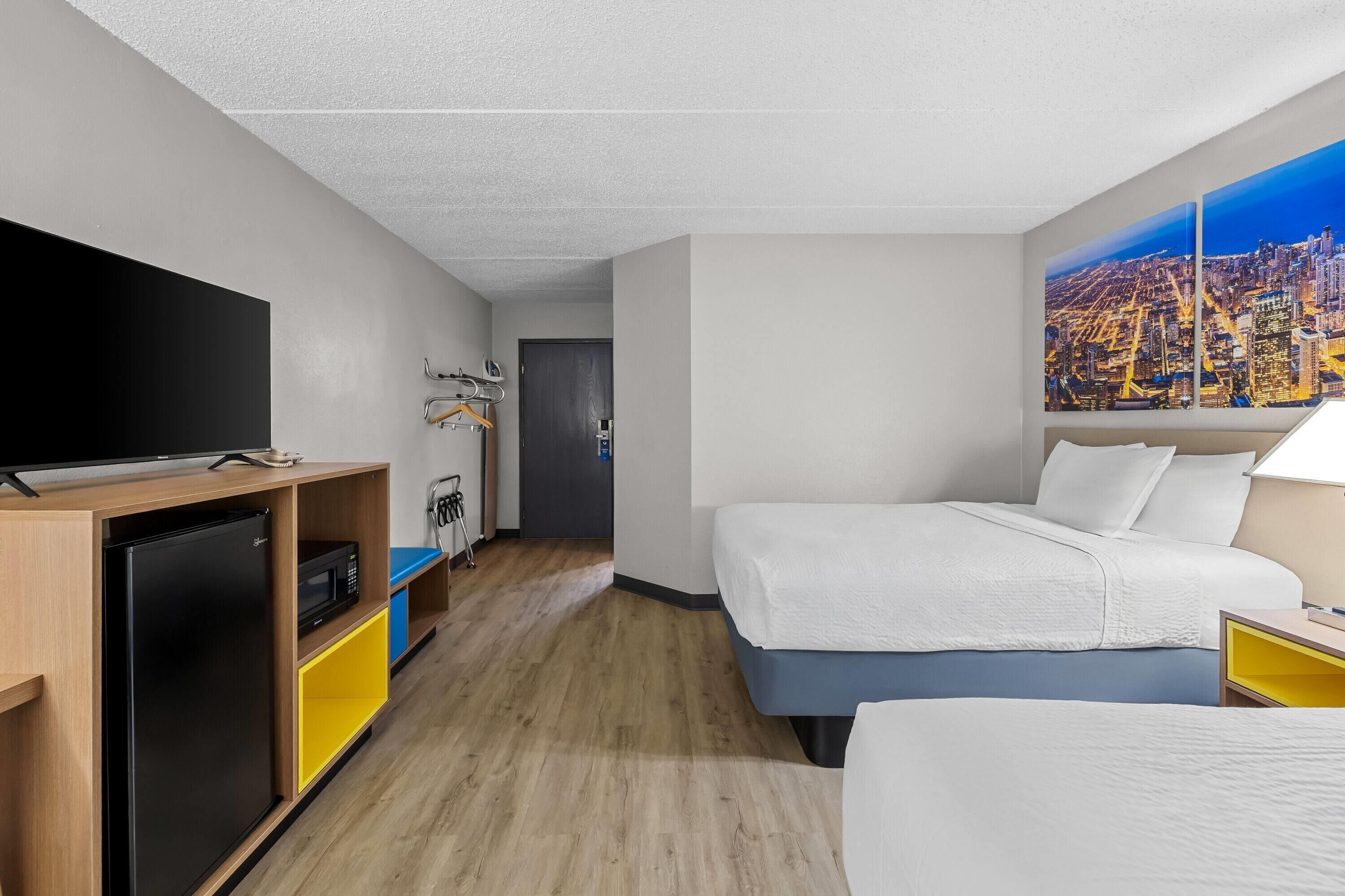 A hotel room with a bed, a TV, a refrigerator, a microwave, a painting of a cityscape, and a door leading to another room.