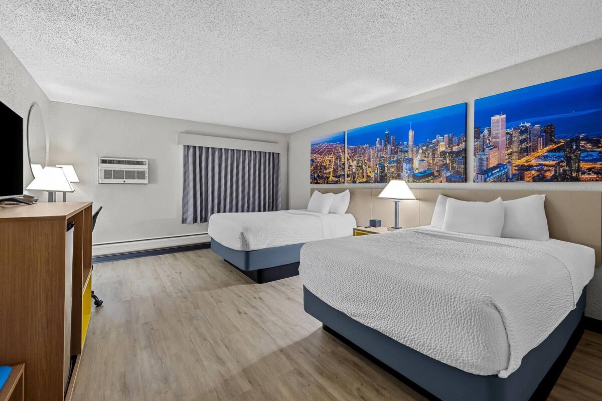 A hotel room with two beds, a desk, a chair, a TV, air conditioning, a window with curtains, and a cityscape painting.