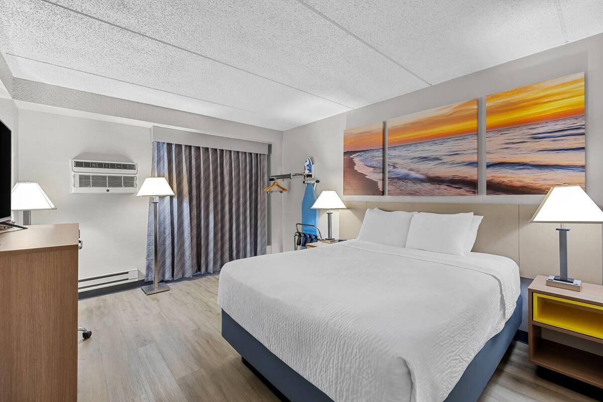 A hotel room with a large bed, two bedside lamps, a desk, a TV, an air conditioner, a curtain, and a sunset painting on the wall.