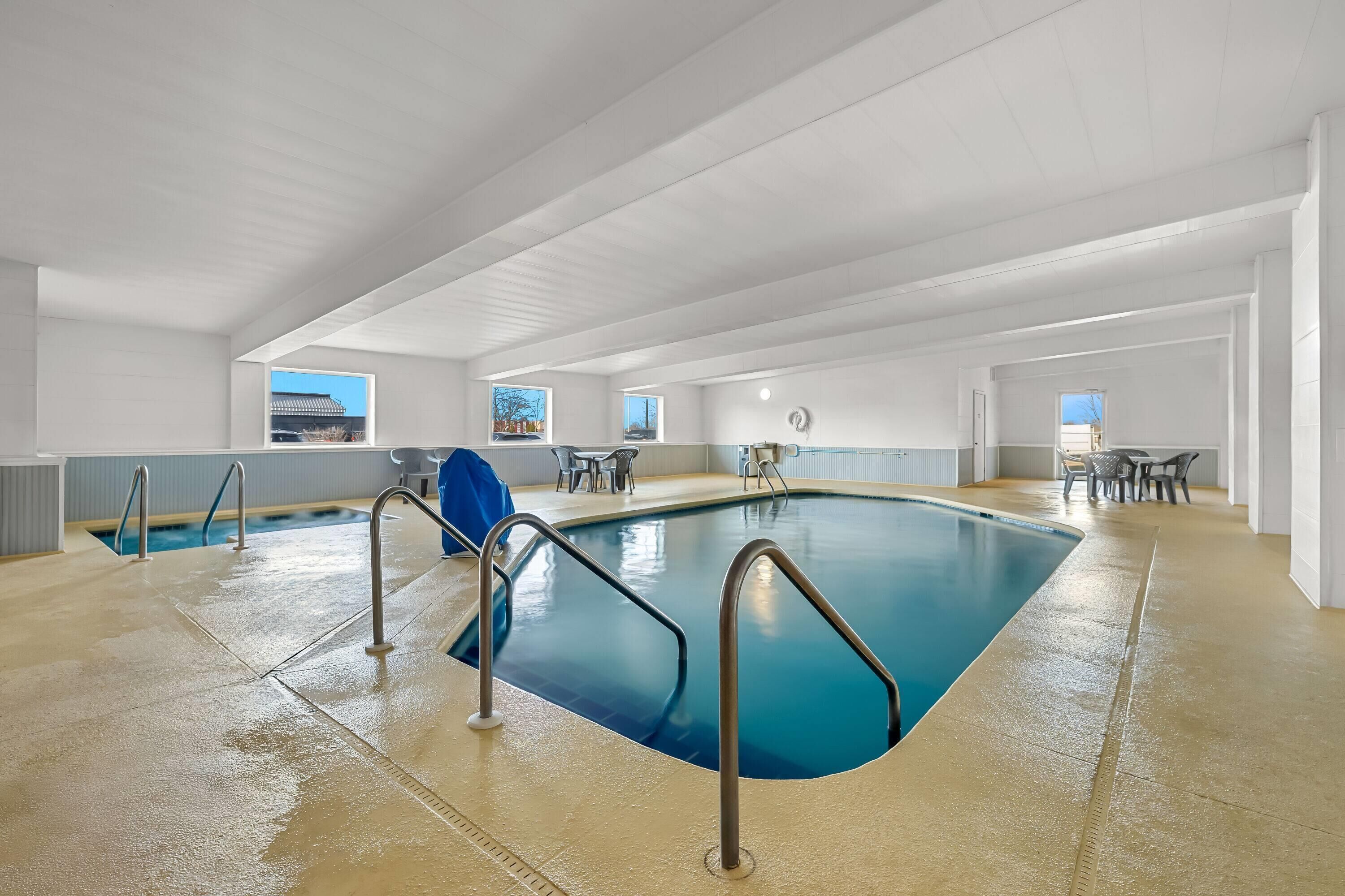 An indoor swimming pool with clear blue water, metal pool ladders, and a view of the outside through windows.