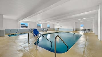 An indoor swimming pool with clear blue water, metal pool ladders, and a view of the outside through windows.