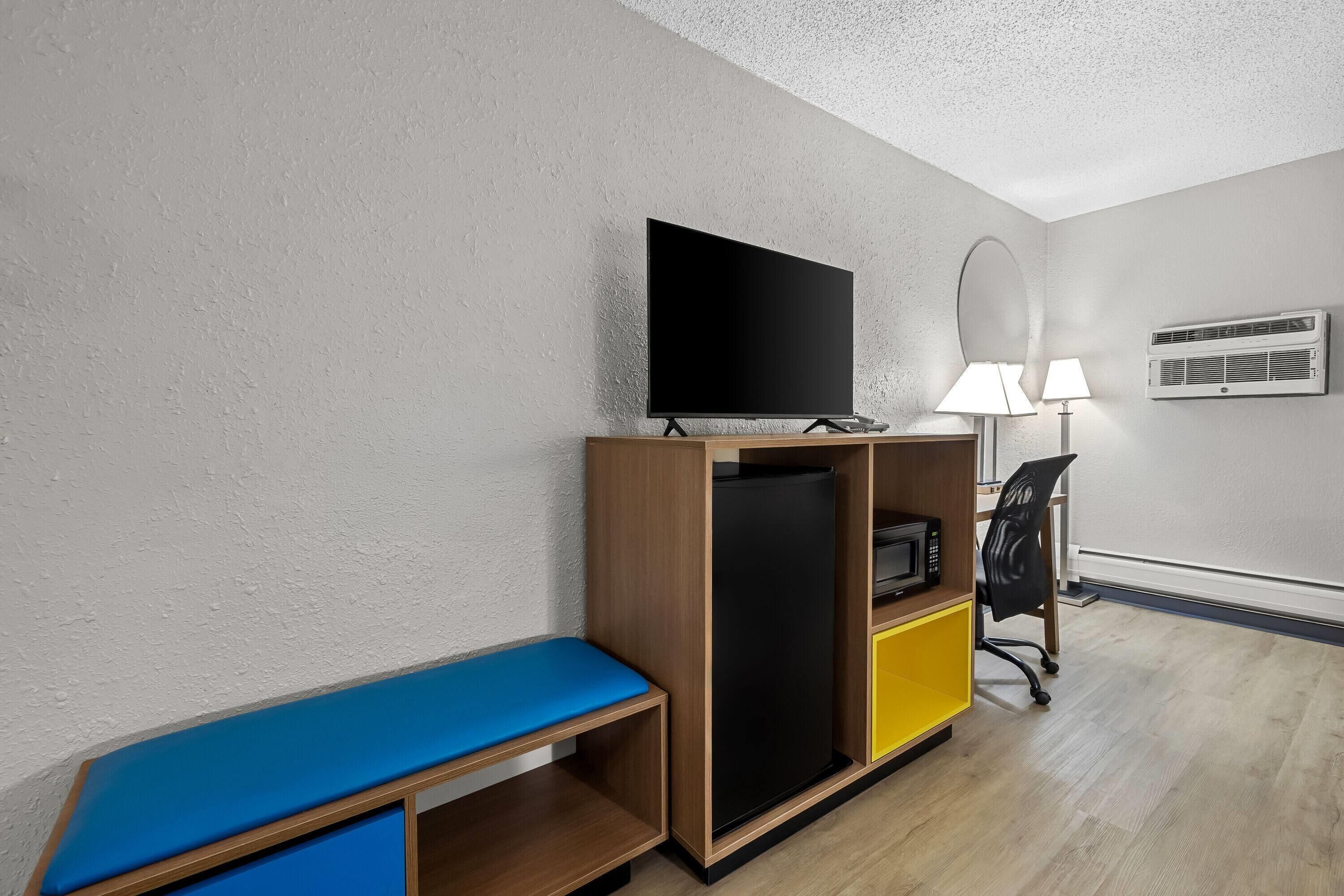 A hotel room with a flat-screen TV, a small refrigerator, a desk with a chair, a bench, and a lamp.