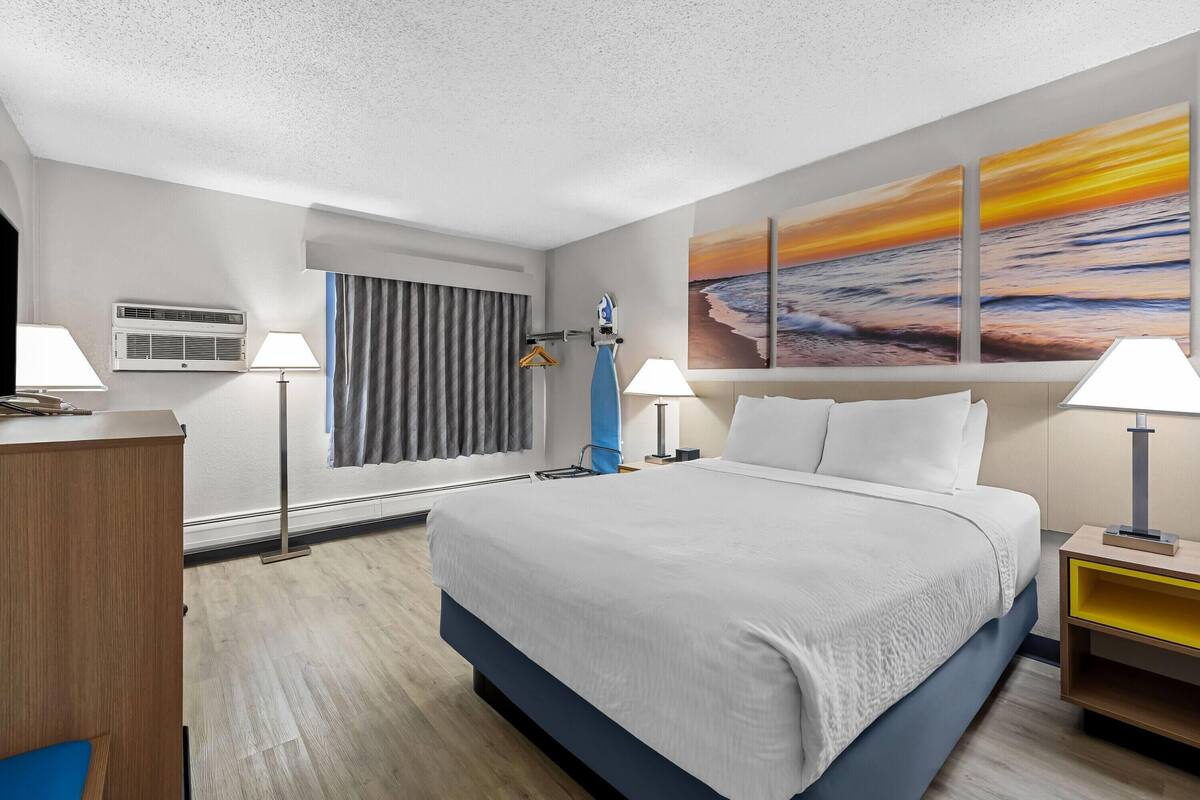 A hotel room with a bed, two bedside lamps, a painting of a sunset, a desk, and a chair.