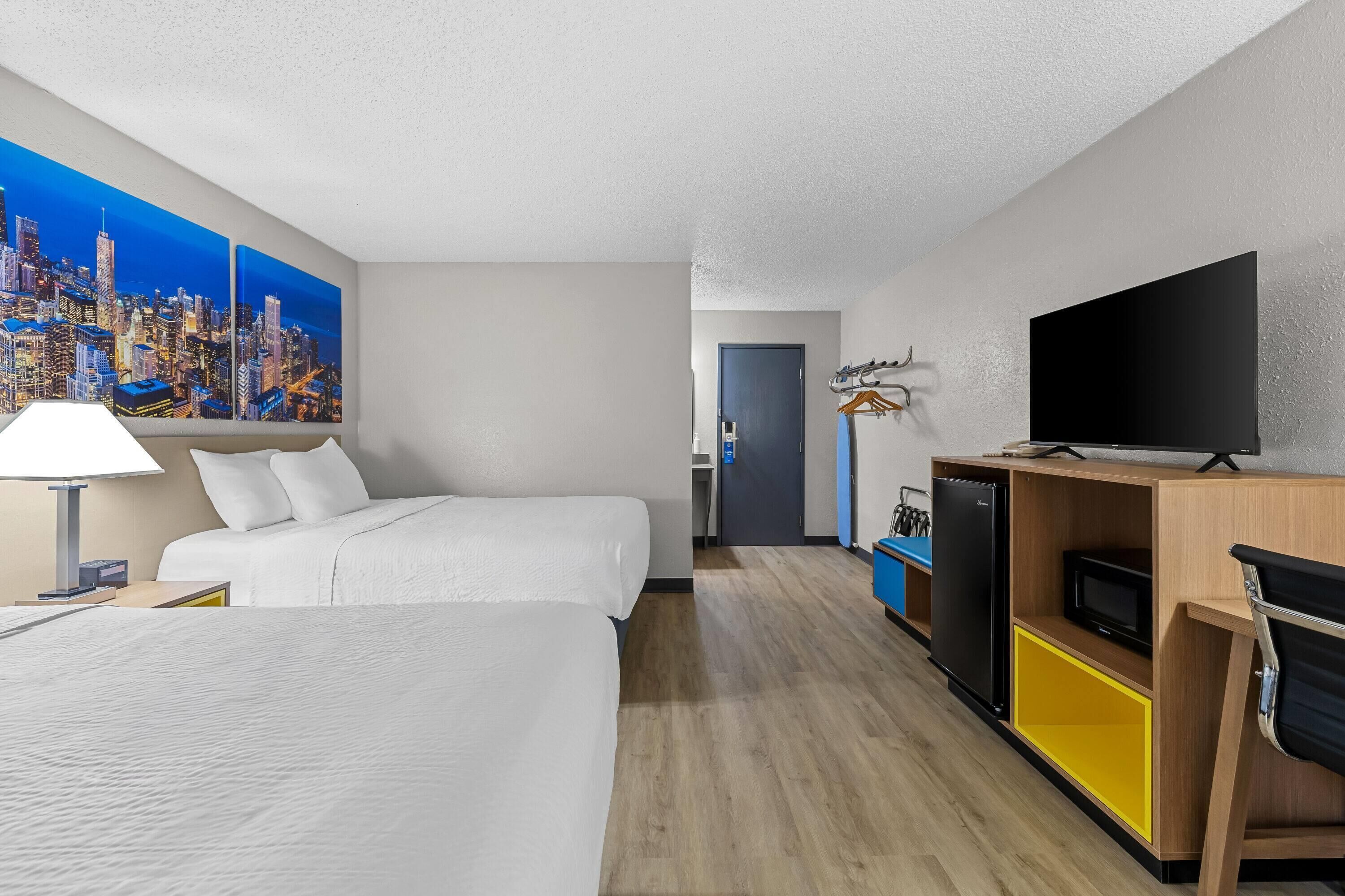 A hotel room with two beds, a television, a desk, and a cityscape painting on the wall.