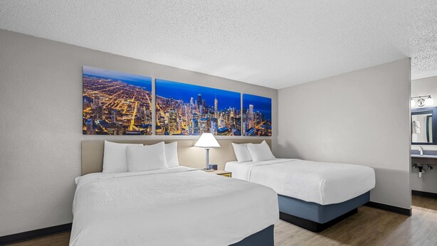 A hotel room with two beds, a cityscape painting, a nightstand, and a bathroom visible. - Days Inn by Wyndham Fond du Lac (Fond du Lac)