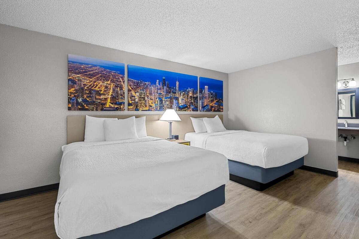 A hotel room with two beds, a cityscape painting, a nightstand, and a bathroom visible.
