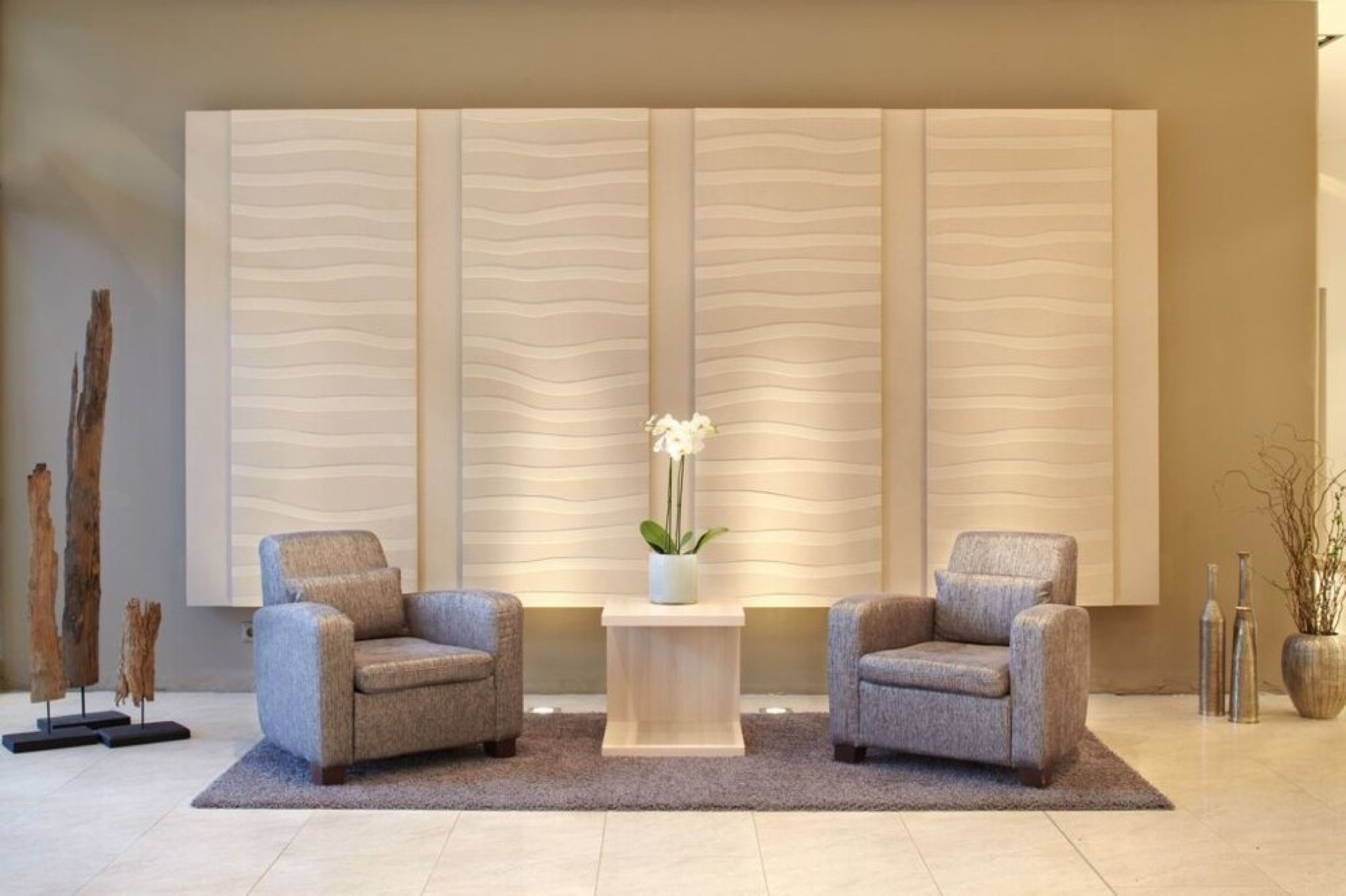 A modern lobby with two grey armchairs, a central table with a vase of flowers, and a textured wall panel.