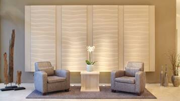 A modern lobby with two grey armchairs, a central table with a vase of flowers, and a textured wall panel.