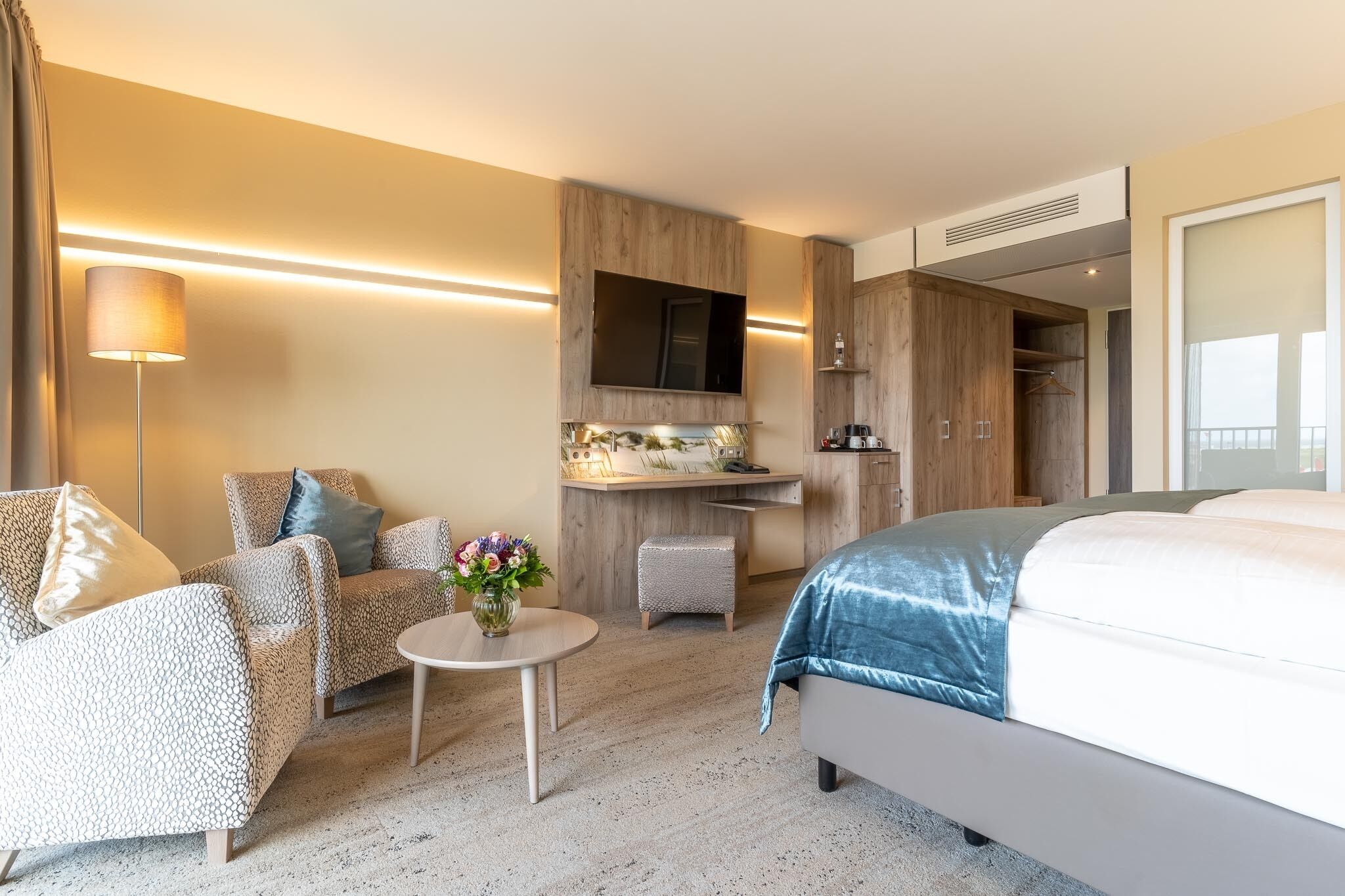 A modern hotel room with a large bed, a seating area with a table and chairs, a kitchenette, and a TV mounted on the wall.