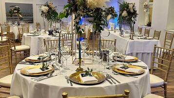 Une salle de banquet avec des tables rondes dressées pour un événement formel, agrémentée de nappes blanches, d’assiettes à bord doré et de grands centres de table floraux.