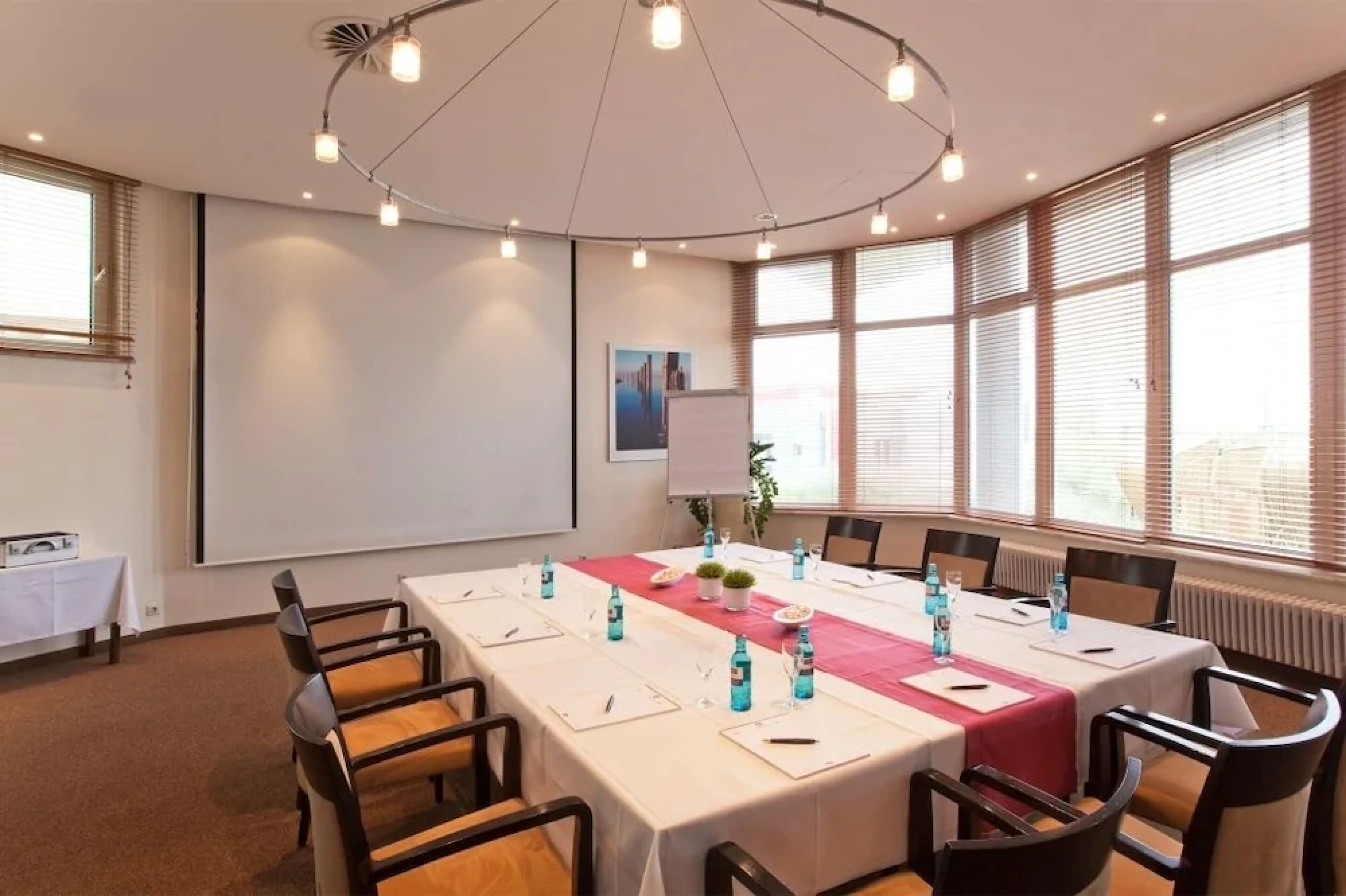 A conference room with a long table, chairs, a projector screen, and a large window with blinds.