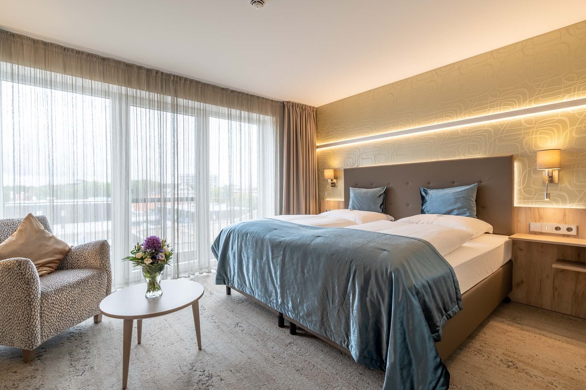 A hotel room with a large bed, a sofa, a small table, and a view of the city through large windows.