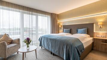 A hotel room with a large bed, a sofa, a small table, and a view of the city through large windows.