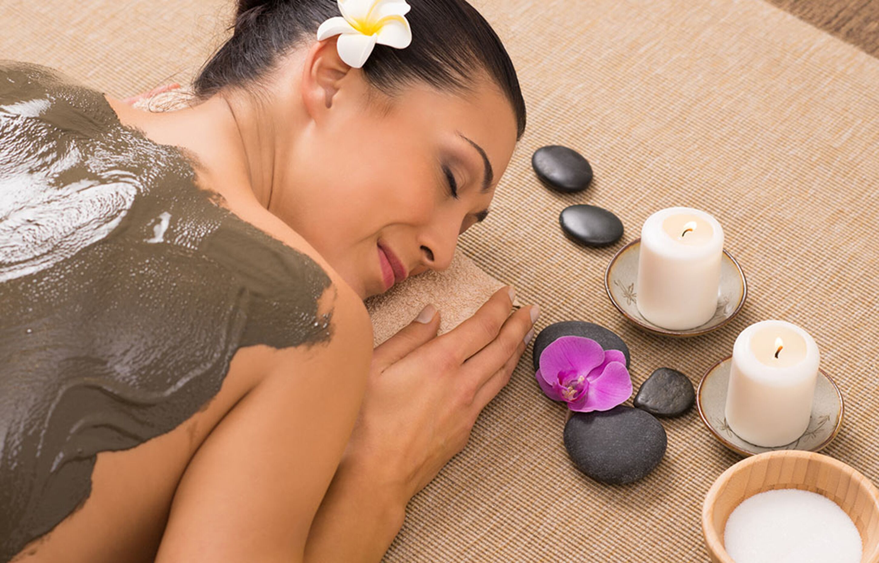 Un ambiente de spa con una persona recibiendo un baño de arcilla, rodeado de piedras, velas y una flor.