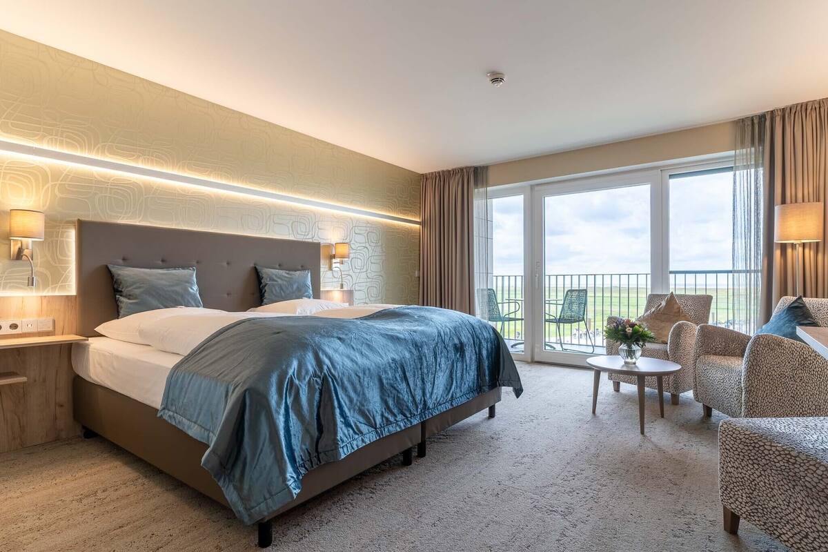 Habitación de hotel con una cama grande, una zona de estar con una silla y una mesita, y un balcón con vistas.