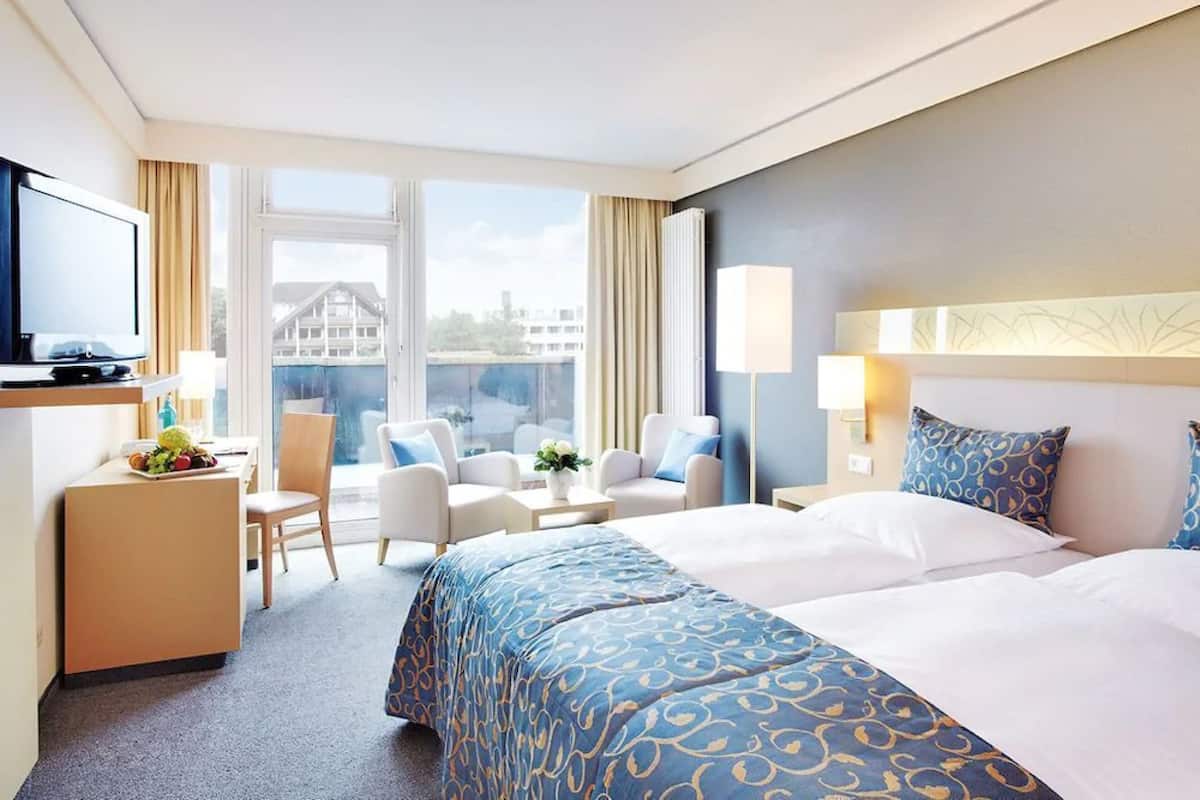 Habitación de hotel con una cama grande, un escritorio con computadora, una silla, una mesita con un jarrón de flores y una ventana con vista a edificios.