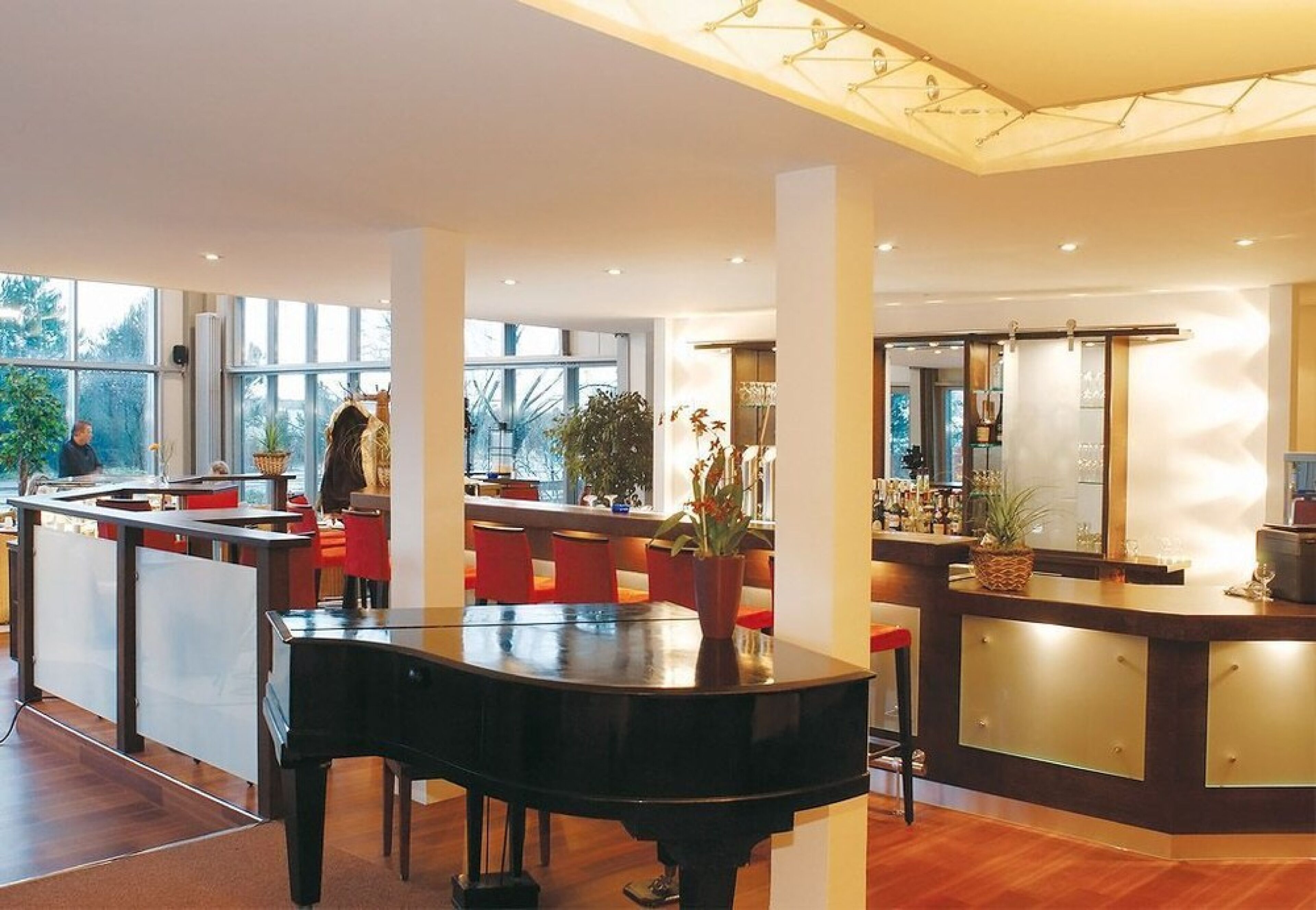 Eine moderne Hotelhalle mit Klavier, roten Sesseln, einer Bar und großen Fenstern.