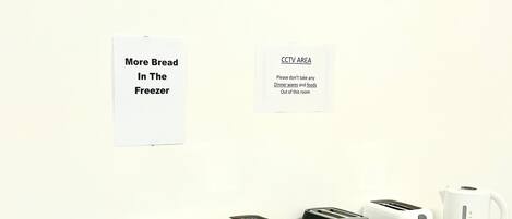 Cocina con cuatro tostadoras, una tetera y recipientes de plástico sobre una encimera. Se observan carteles en la pared que indican una zona de videovigilancia y más pan en el congelador.