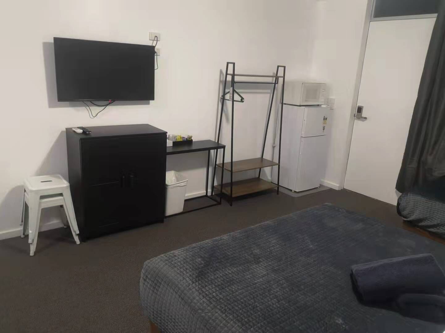 A bedroom with a bed, a desk with a chair, a television, a wardrobe, and a small stool.