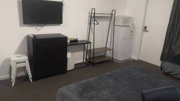 A bedroom with a bed, a desk with a chair, a television, a wardrobe, and a small stool.