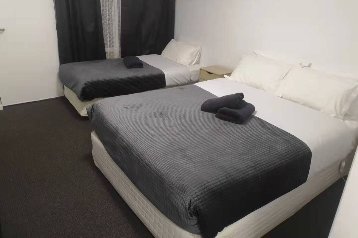 A hotel room with two beds, each with a grey bedspread and white pillows. There is a nightstand with a lamp between the beds.
