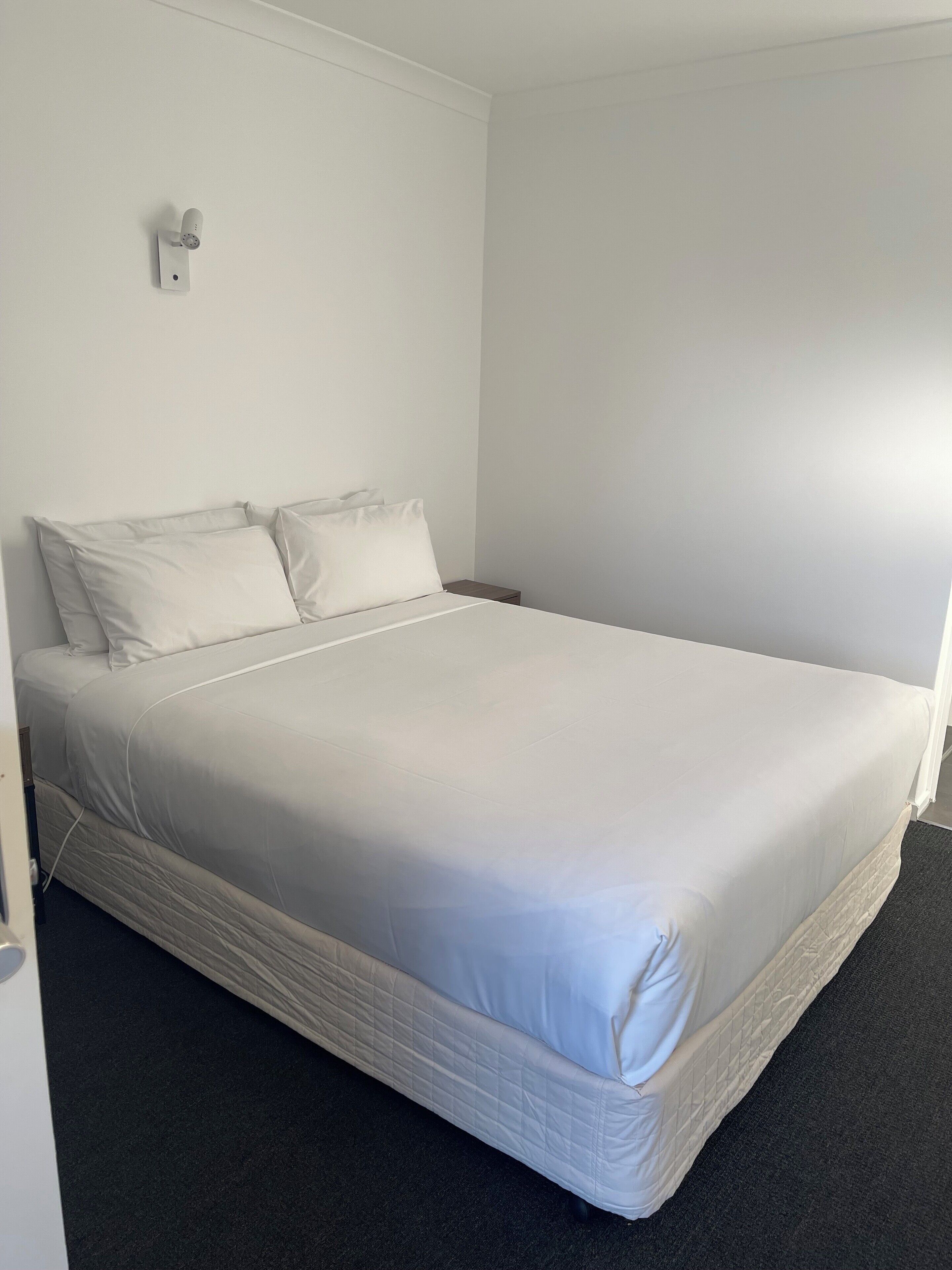 A single-bed room with a white mattress, two pillows, a bedside table, and a wall-mounted light fixture.
