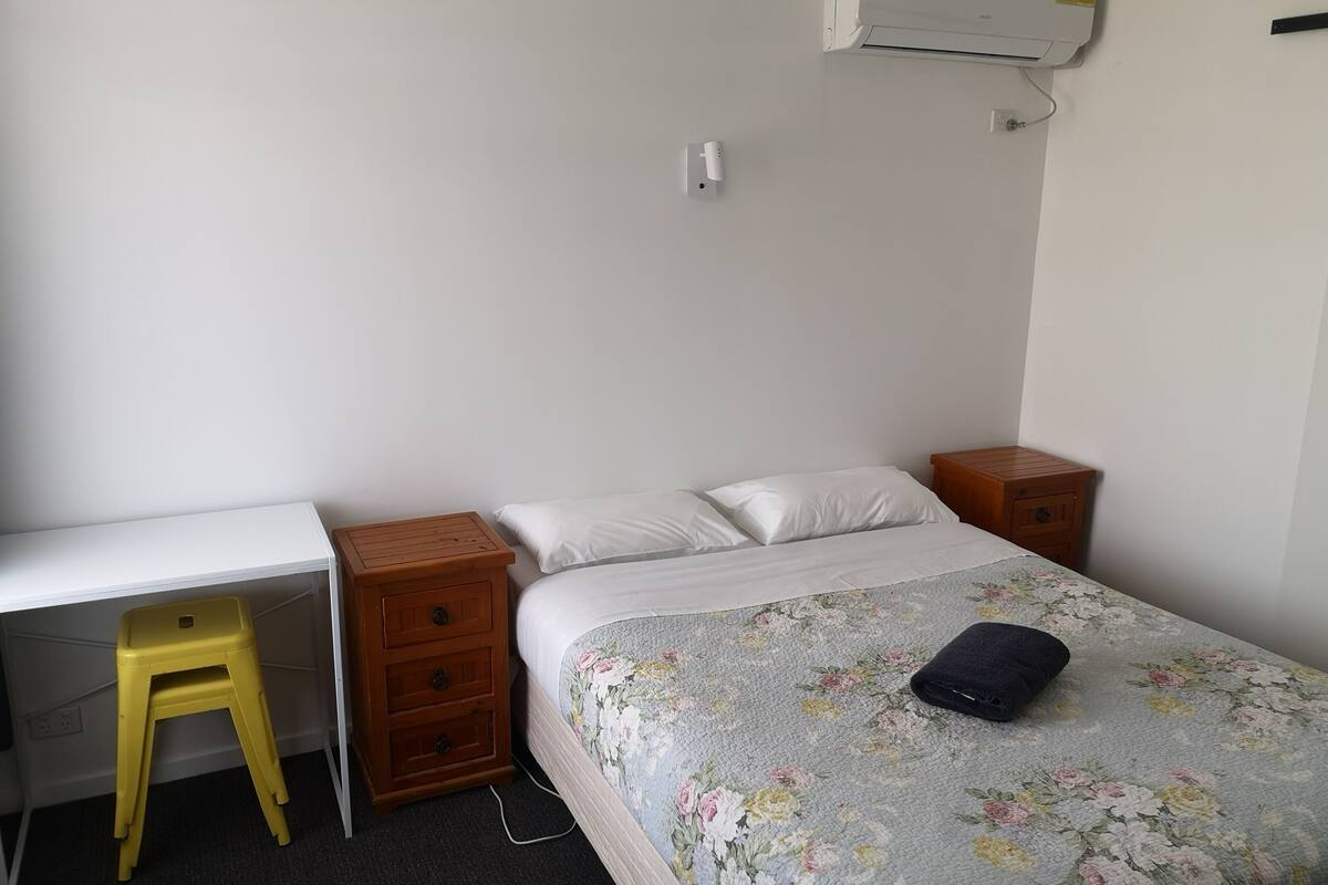 A bedroom with a bed, a desk, a chair, and a small wooden nightstand.