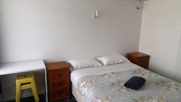 Quarto com cama, escrivaninha, cadeira e uma pequena mesa de cabeceira de madeira.
