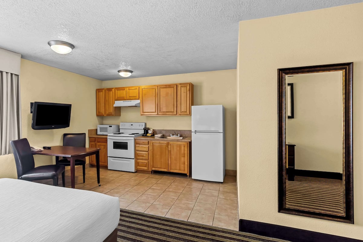 A compact hotel room with a kitchenette, a dining area, a flat-screen TV, and a large mirror.