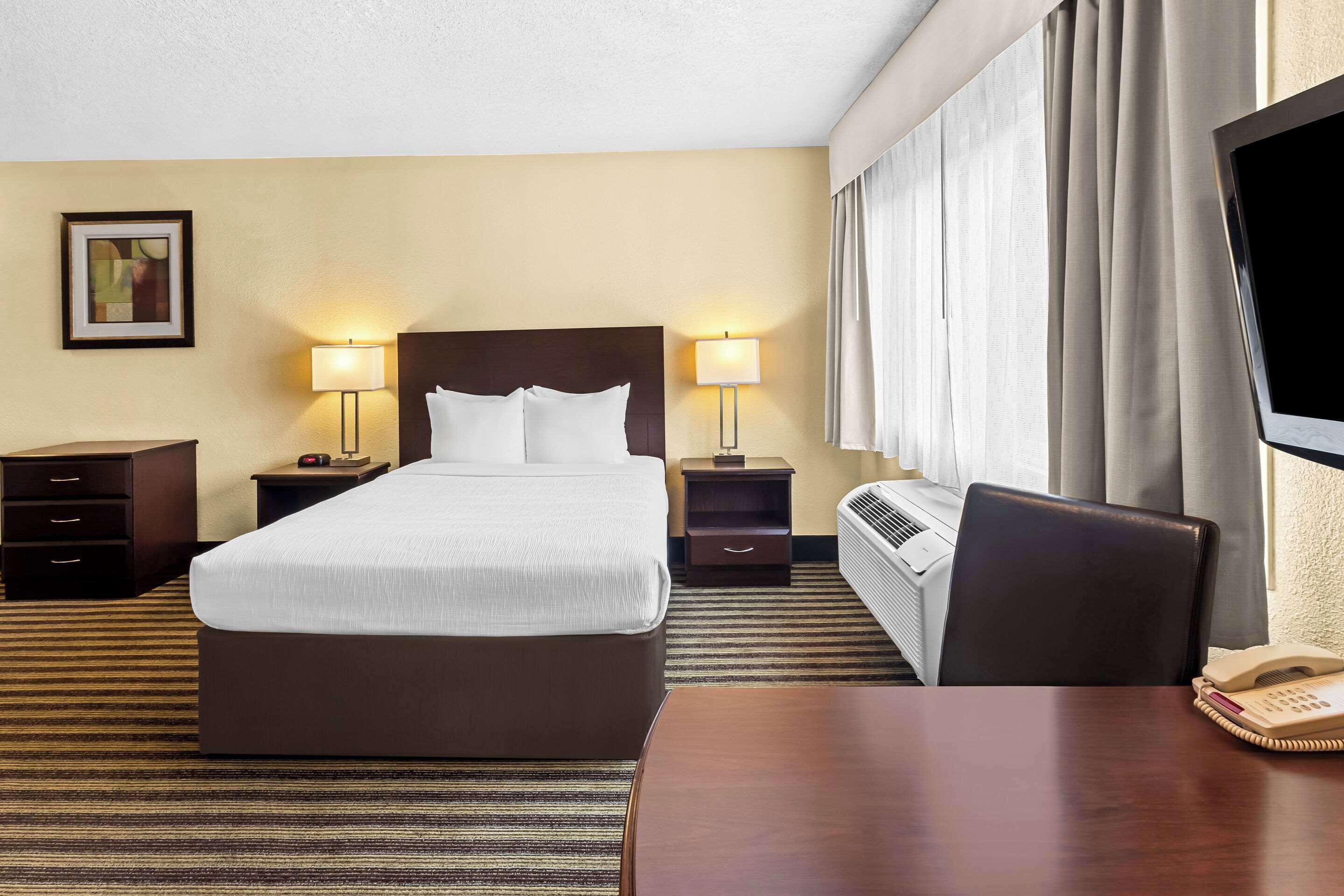 Habitación de hotel con una cama grande, dos mesitas de noche con lámparas, un escritorio con silla, un televisor y una ventana con cortinas.
