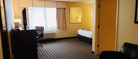 A hotel room with a bed, a desk with a computer, a chair, a trash bin, and a window with curtains.