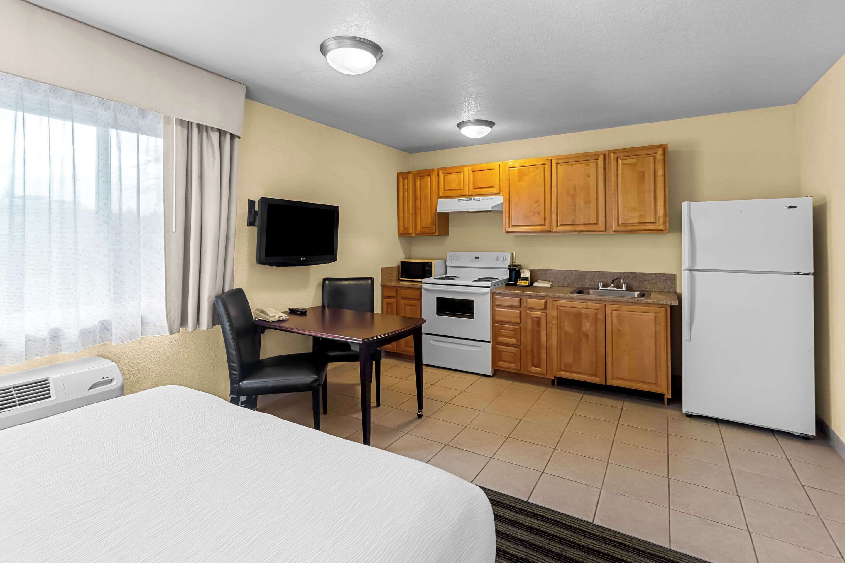 A compact hotel room with a bed, a dining table, a TV, a kitchenette with a stove, microwave, and refrigerator.