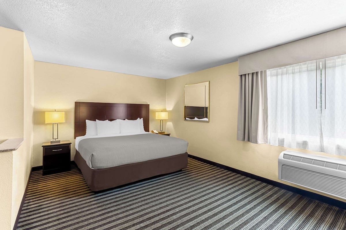 A hotel room with a large bed, two bedside tables with lamps, a mirror, a window with curtains, and a wall-mounted air conditioning unit.