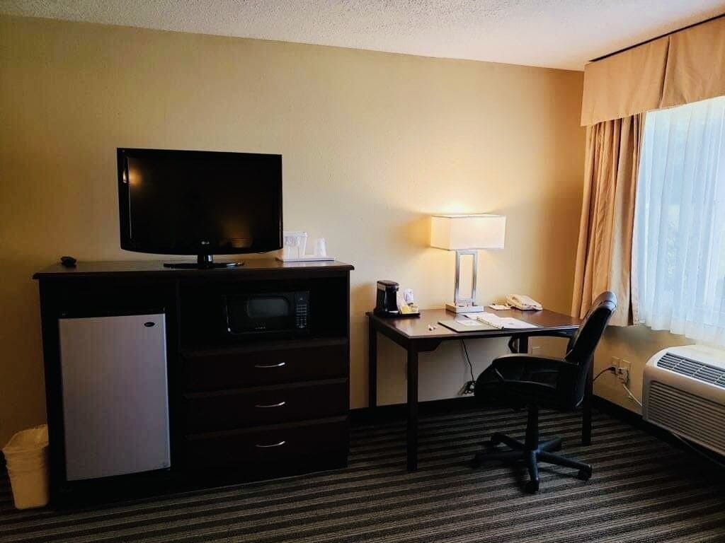 A hotel room with a flat-screen TV, a mini-fridge, a desk with a lamp, a chair, and a window with curtains.