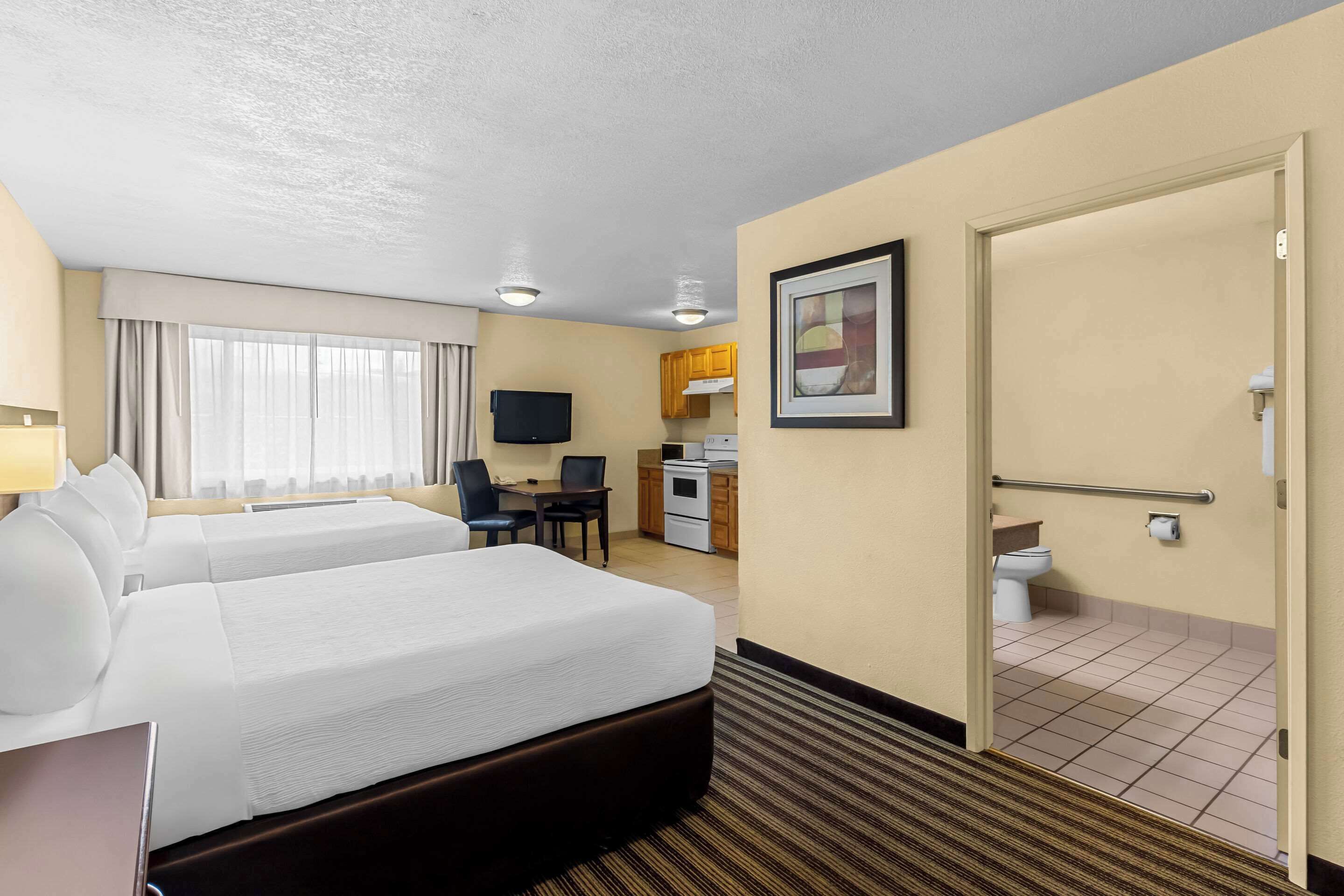 A hotel room with two beds, a desk, a television, a kitchenette, and a bathroom.